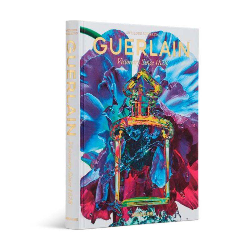 Livro Guerlain Visionary Since 1828 da Assouline com capa dura sobre mesa de centro de madeira nobre ao lado de um frasco de