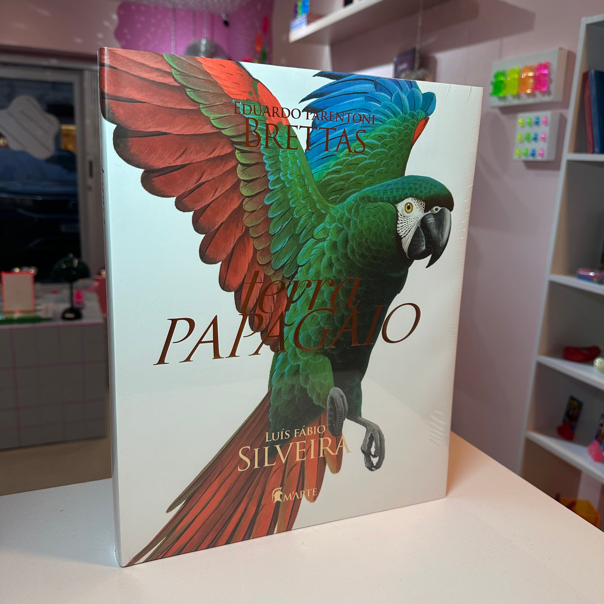 Livro Terra Papagaio com capa dura colorida sobre mesa de centro de madeira em sala de estar decorada com plantas tropicais e