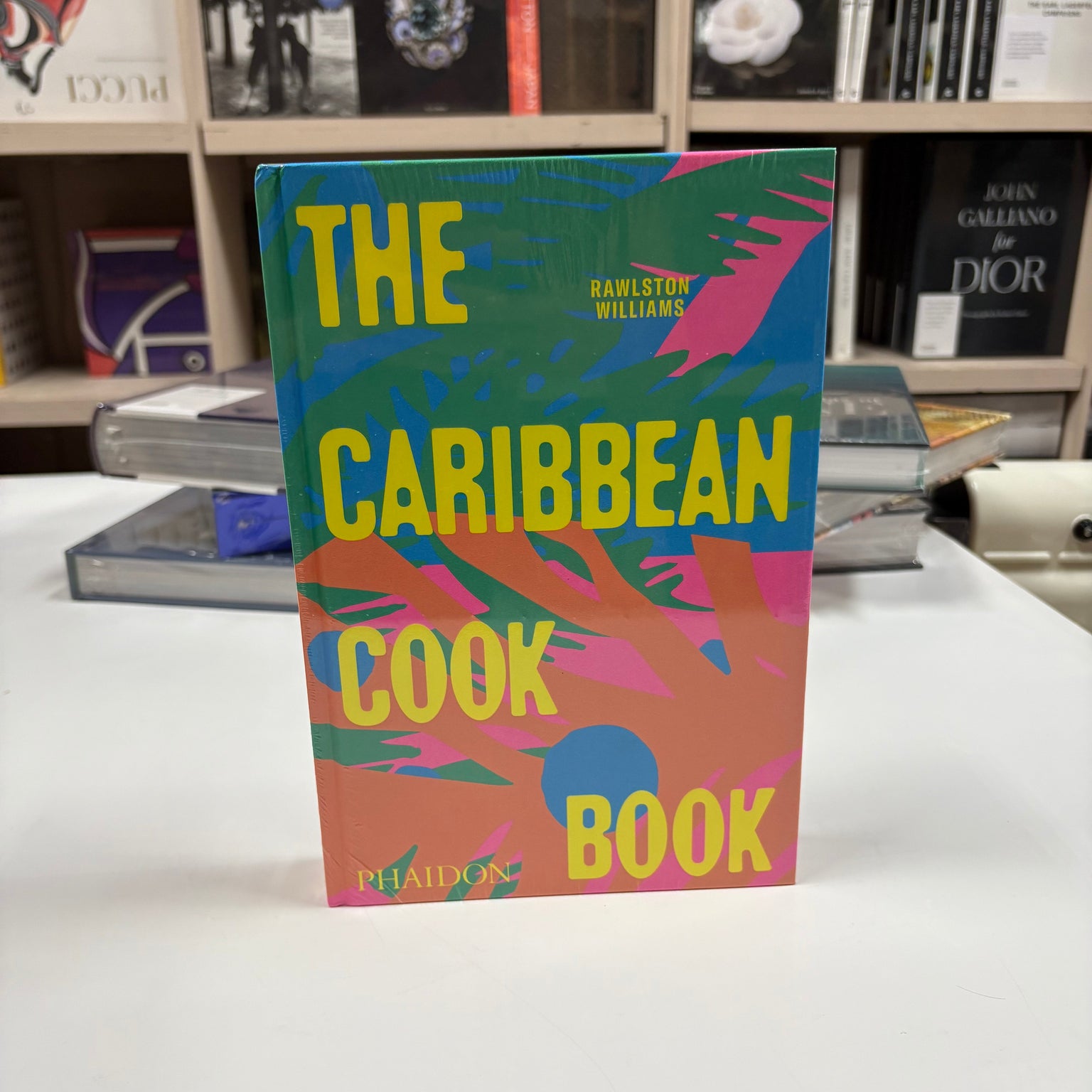 Livro The Caribbean Cookbook Phaidon Press com capa colorida sobre mesa de centro de madeira ao lado de frutas tropicais e um