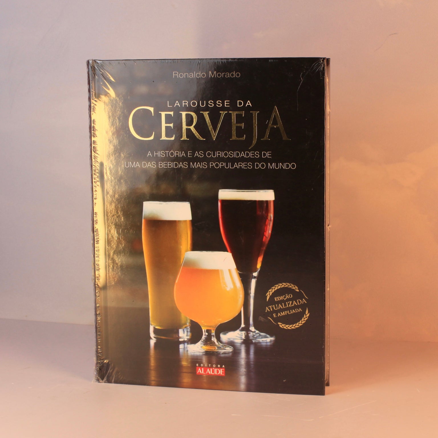 Livro Larousse da Cerveja com capa dura sobre mesa de centro de madeira ao lado de um copo de cerveja artesanal e lúpulo, em