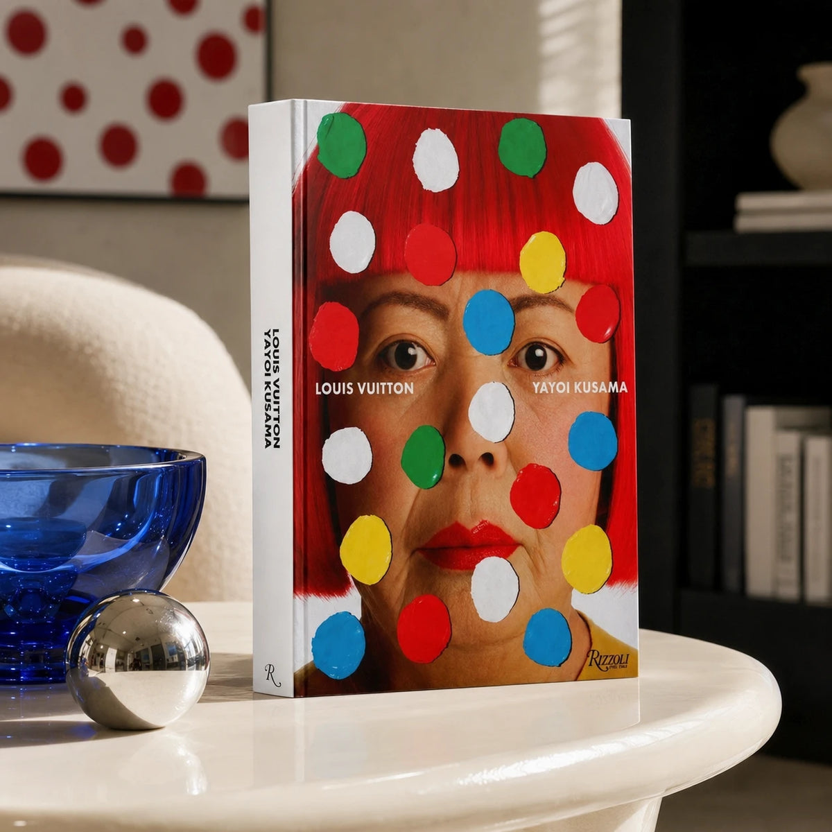 Livro Louis Vuitton Yayoi Kusama Rizzoli com capa dura colorida sobre mesa de centro de mármore ao lado de uma escultura cont