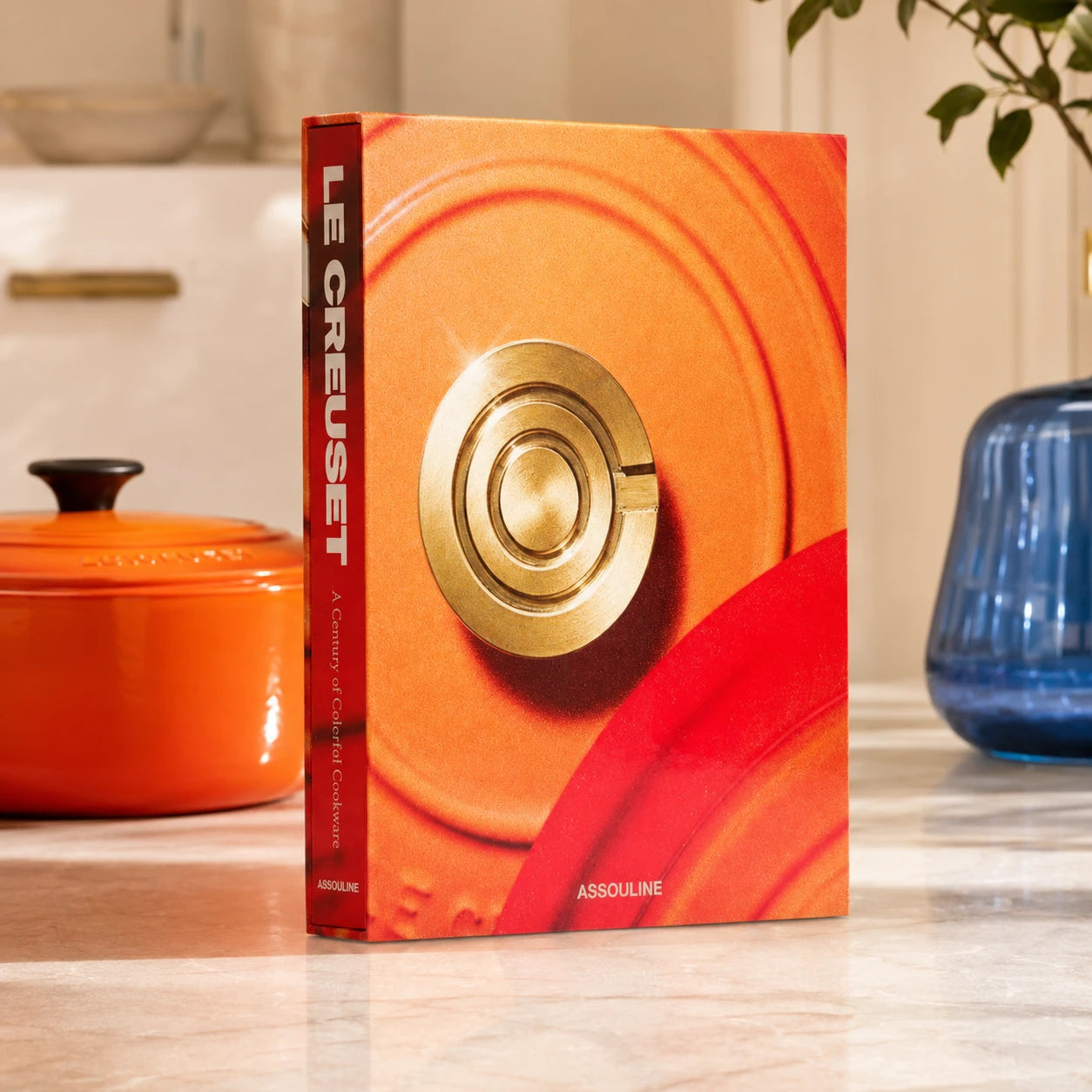 Livro Le Creuset Assouline com capa dura colorida sobre mesa de centro em cozinha gourmet iluminada, ao lado de peças da marc